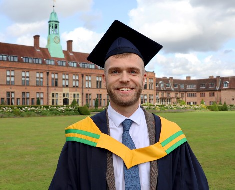 Colin Hartley wearing his graduation cap and gown over a shirt and tie stands on the lawn in front of the HCA Building.